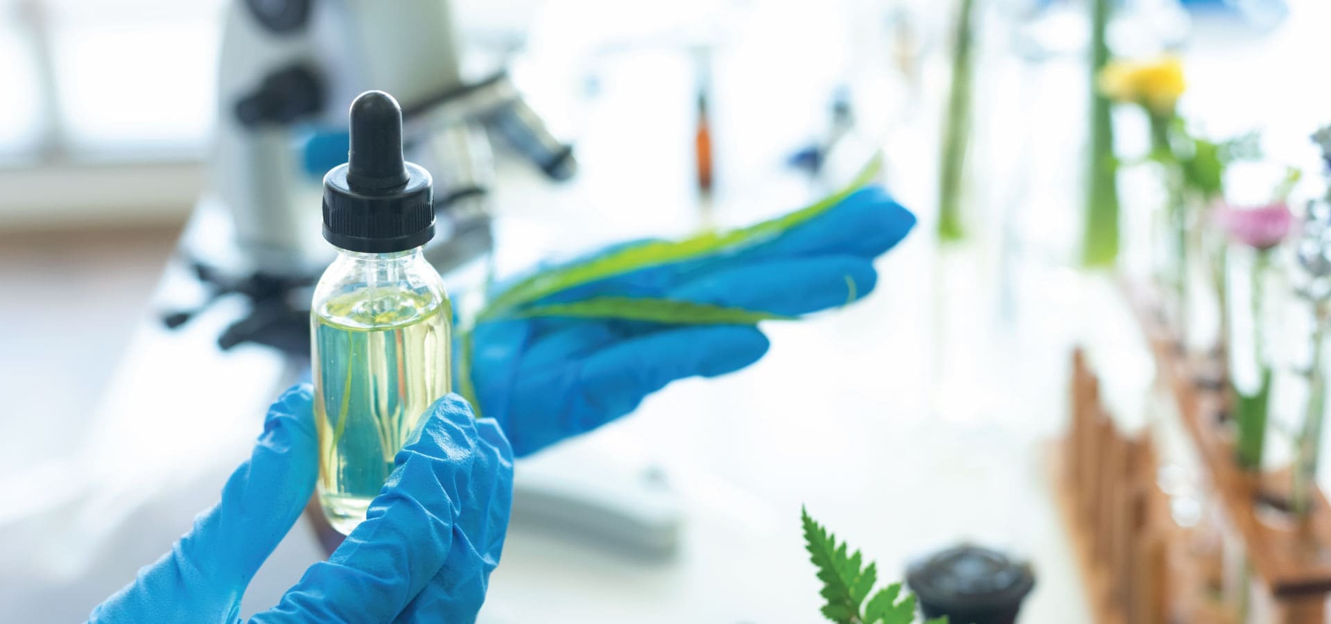 A researcher in blue gloves holds a dropper bottle of golden liquid, with a green leaf, amidst lab equipment and plants in the background.