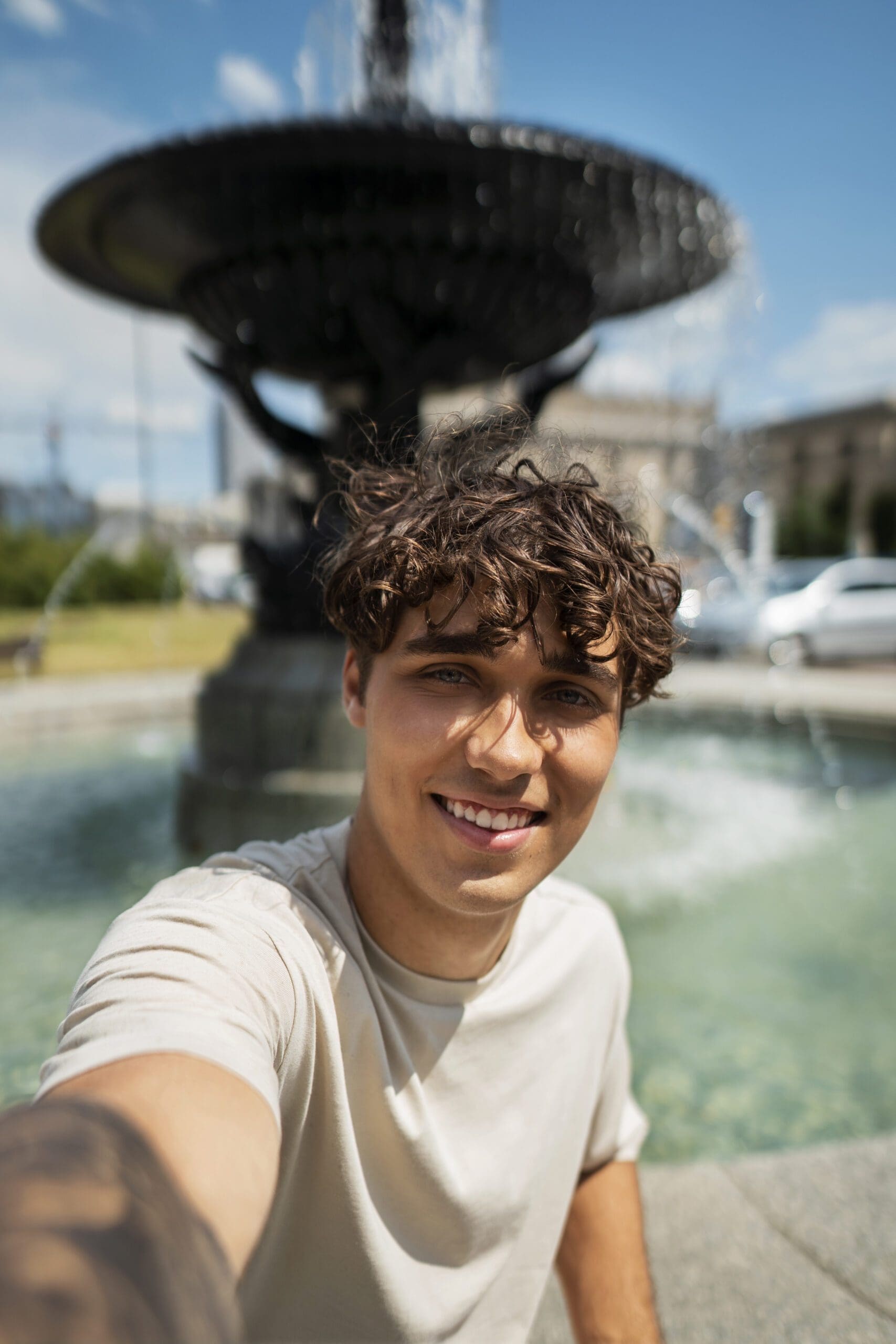 A man takes a selfie in front of a fountain, surrounded by greenery and a clear blue sky. Water gently splashes in the background.