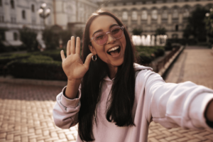 A beautiful woman in a pink hoodie stands in a garden, waving with one hand, surrounded by greenery and elegant architecture in the background.