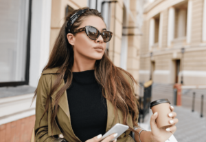 A woman in stylish attire holds a smartphone and coffee cup, standing on a city sidewalk near elegant architecture.