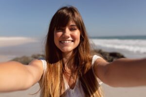 A beautiful woman with long, wavy hair takes a selfie on a sunny beach, with waves and a clear blue sky in the background.