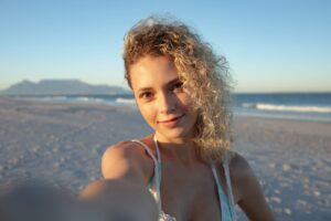 A woman with curly hair stands on a beach, holding a camera and smiling, with ocean waves and mountains in the background.