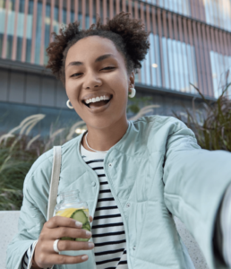 A woman with curly hair wears a light blue jacket and striped shirt, holding a drink with lime in a jar, posing outdoors.