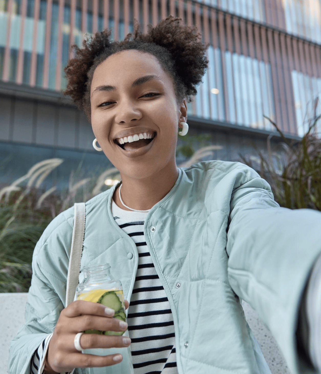 A woman with curly hair wears a light blue jacket and striped shirt, holding a drink with lime in a jar, posing outdoors.
