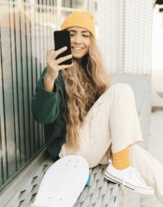 A woman sits on a bench, wearing a green sweater, yellow cap, and white sneakers, holding a phone and resting on a skateboard.