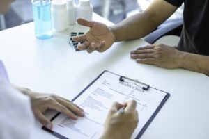 A person fills out a health check form on a clipboard while another gestures across the table, medicine bottles in the background—raising questions like, “Does CBD show up on a drug test?”.