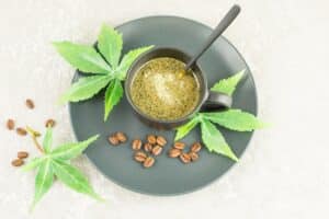 A cup of dark liquid with a spoon, surrounded by green leaves and coffee beans on a gray plate, highlights the benefits of including hemp in diet for a nutritious boost.