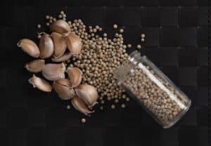 Garlic cloves and white peppercorns, perfect for adding flavor and boosting the benefits of hemp in your diet, are scattered on a black woven surface beside an open glass jar lying on its side.