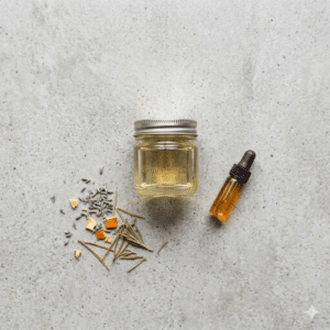 A small glass jar of clear liquid, a dropper bottle with amber liquid, and a small pile of dried herbs and orange peel on a light stone surface.