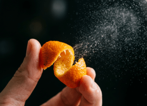 A hand squeezes an orange peel, spraying a fine mist of citrus oil into the air against a dark background.