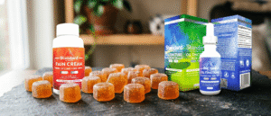 Bottles of pain cream and oil tincture, along with their boxes, are displayed on a table with several orange gummies arranged in front.