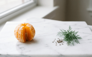 A peeled orange, a small pile of seeds, and a sprig of dill on a white marble surface near a window.