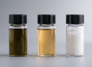 Three small glass jars with black lids, containing a dark liquid, a light yellow liquid, and white crystalline granules, are arranged side by side on a neutral background.