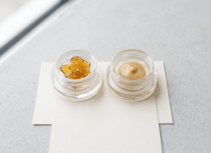 Two small glass jars sit side by side; the left jar contains a translucent amber substance, while the right jar holds a creamy beige paste. Both are placed on white paper atop a gray surface.