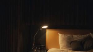 A dimly lit bedroom with a bedside lamp glowing beside white pillows at night