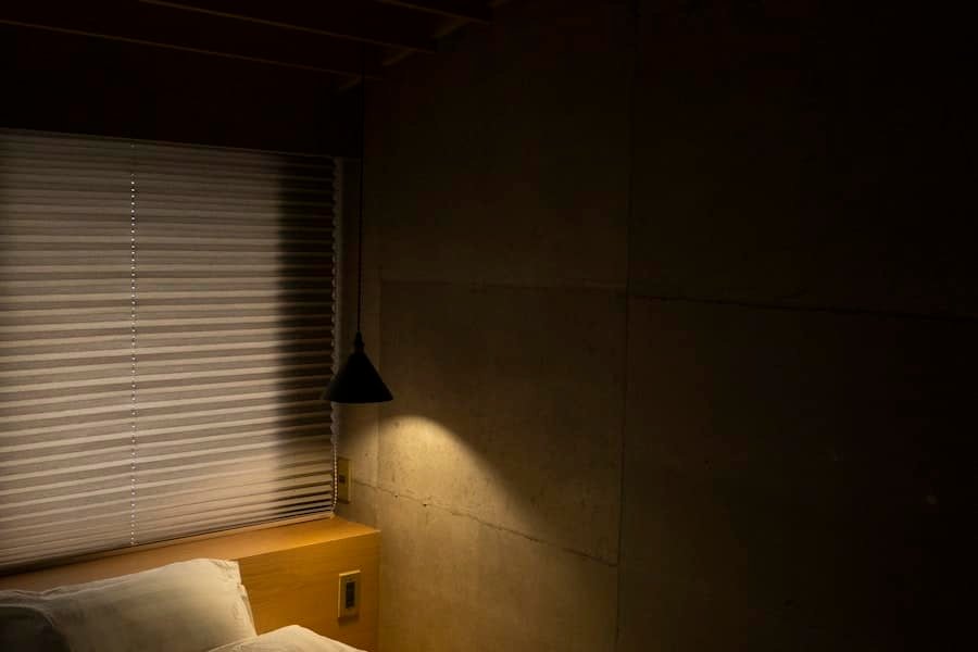 A darkened bedroom with window blinds and a wall lamp above the bed