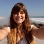 A beautiful woman with long, wavy hair takes a selfie on a sunny beach, with waves and a clear blue sky in the background.