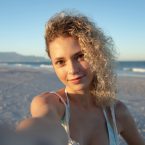 A woman with curly hair stands on a beach, holding a camera and smiling, with ocean waves and mountains in the background.